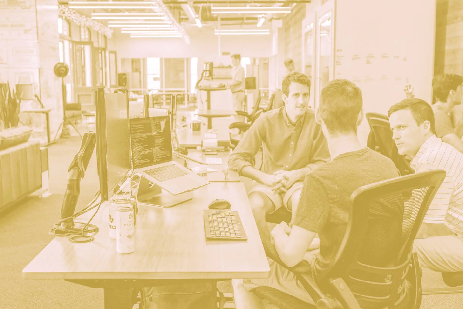 A modern open-plan office where a group of young men are engaged in a collaborative discussion around a desk with multiple computer monitors and a laptop. The laptop screen displays lines of code, suggesting a tech start-up or software development setting. The workspace features ergonomic chairs, light wooden furniture, and a relaxed seating area in the background. Energy drink cans and tech accessories are placed on the desk. Large windows allow natural light to fill the space. The entire image has a warm yellow-tinted filter, giving it a stylised appearance.